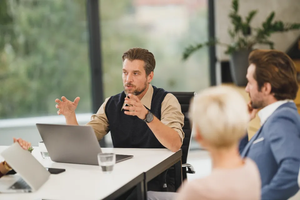 A team lead explaining how his team works to two colleagues across a meeting table with laptops open